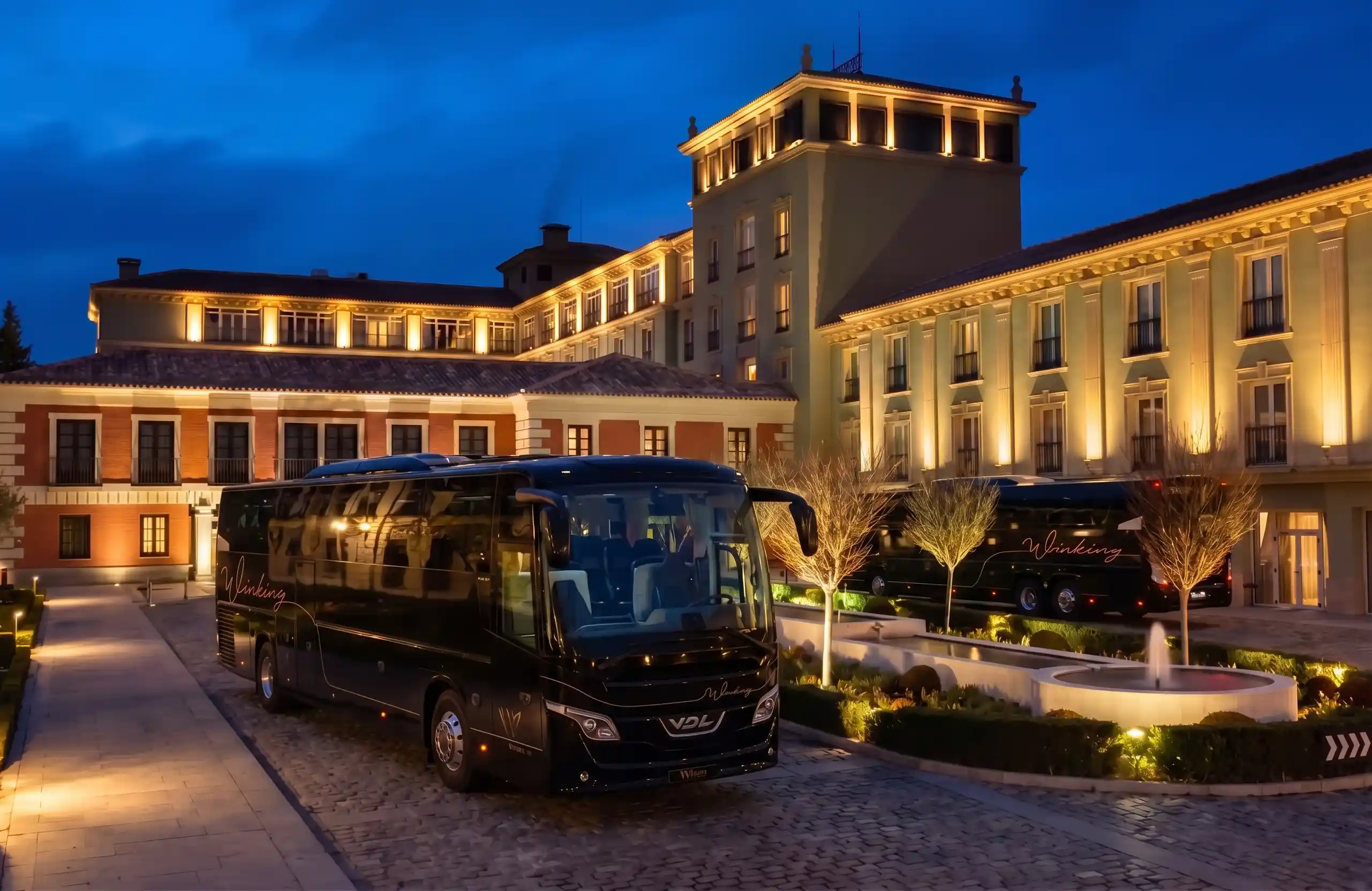 Autocar VDL negro de Winking Bus aparcado en un patio adoquinado ante la fachada iluminada de un hotel, de noche, con una fuente y un segundo autocar al fondo