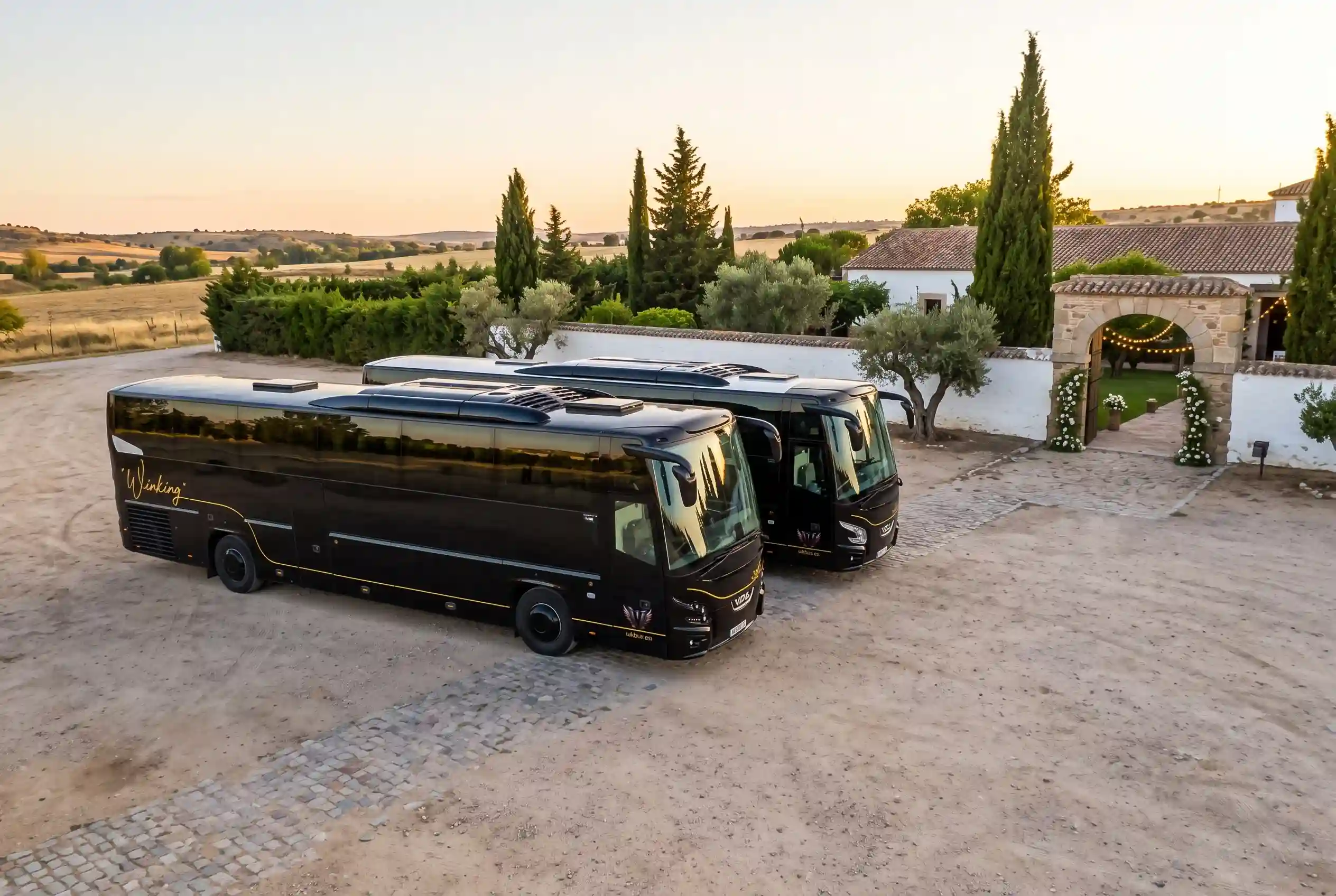 Dos autocares negros de Winking Bus con el rótulo dorado, aparcados en la explanada de una finca de bodas con muro blanco, olivos, cipreses y un arco de entrada con luces al atardecer